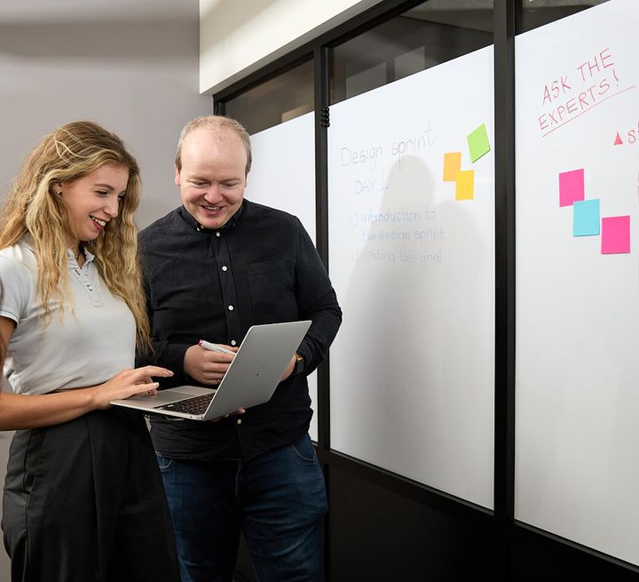 Two team members smiling at a laptop screen in front of a whiteboard